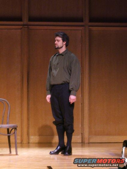 Man in historical costume stands on a wooden stage with a chair to his left