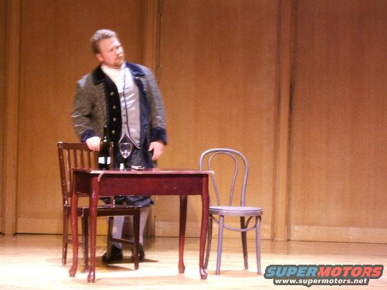 Man in a period costume stands by a table with wine and a glass on a stage
