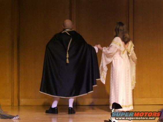 Two people in historical costumes stand on a wooden stage, facing away from the camera