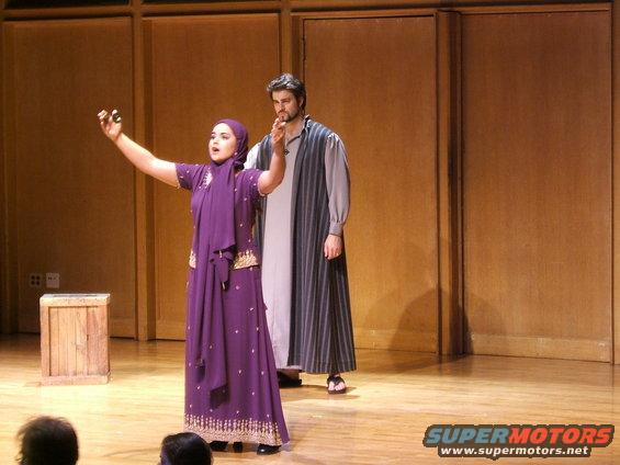 Two actors on a stage, one in a purple outfit and headscarf, the other in a striped robe
