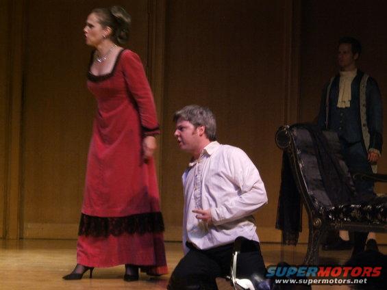Stage actors in period costumes, one in a red dress, another kneeling in white, and a third standing in blue