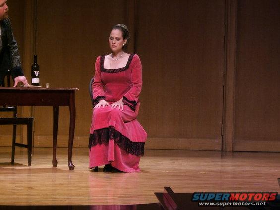 Woman in pink velvet dress with black lace trim sits on stage