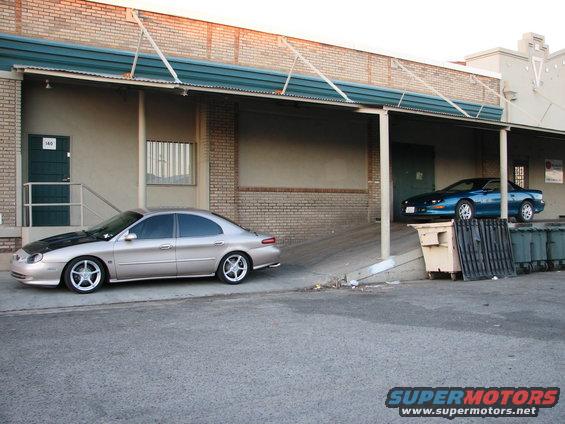 tan sedan and blue coupe parked by a building with dumpsters