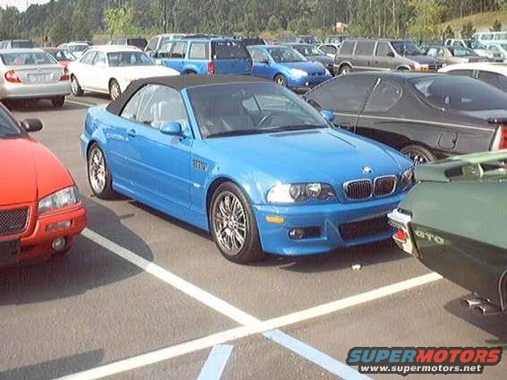 Blue convertible BMW M3 parked in a lot, front view. Red car to the left, green car to the right