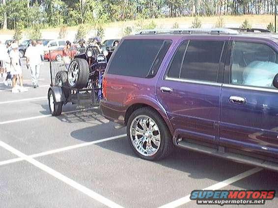 Purple SUV with chrome wheels towing a trailer carrying two spare tires. Other vehicles and people are visible in the background