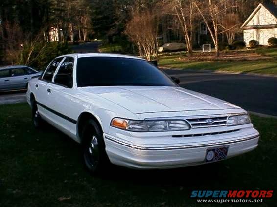 White sedan parked on grass, another car in background. House and trees behind