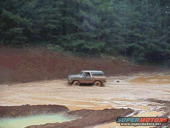 1978 Ford Bronco driving through a muddy area with trees in the background