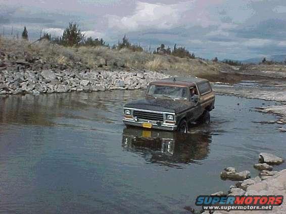 1978 Ford Bronco driving through a shallow body of water with rocky banks