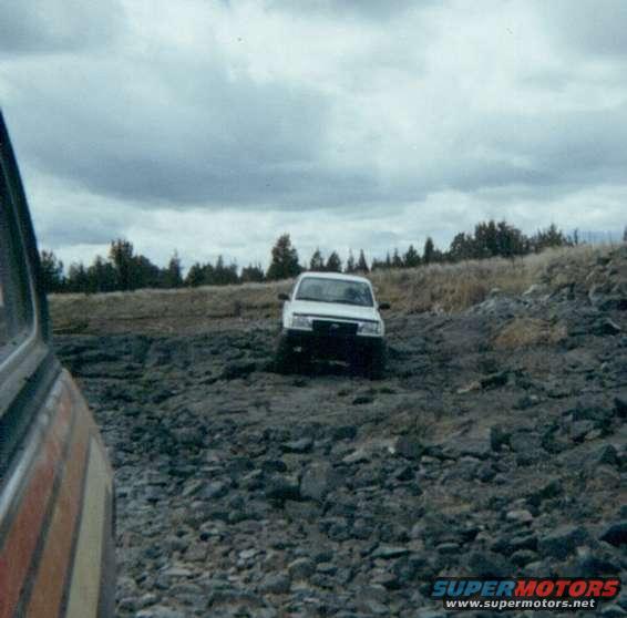 White SUV on rocky ground, with another vehicle's side visible in foreground