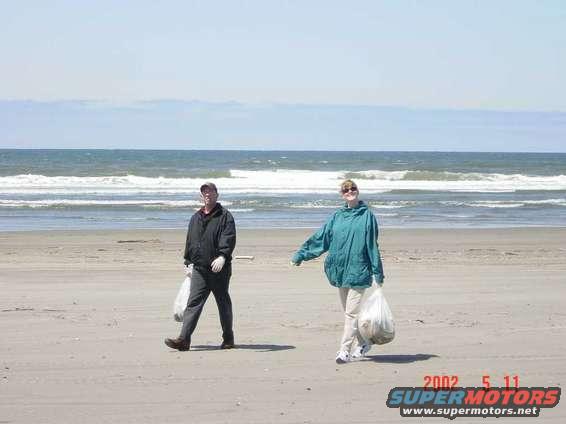 Two people walk on a beach carrying trash bags, with the ocean in the background