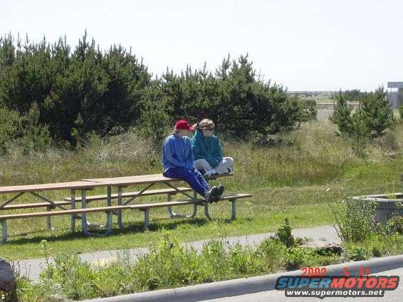 Two people sit on a picnic table near trees and grass. A watermark reads '2002 SUPERMOTORS'
