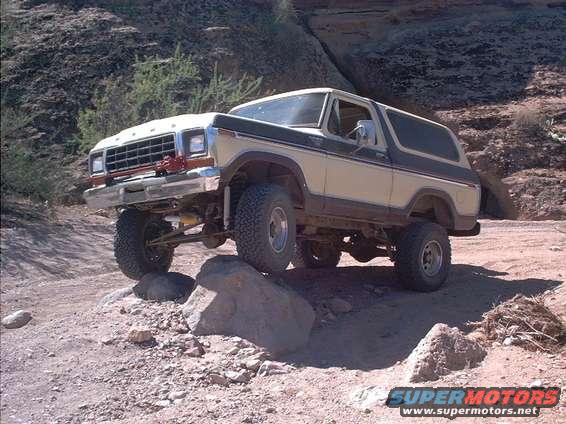 Tan and black 1978 Ford Bronco off-roading over rocks, front-left tire elevated