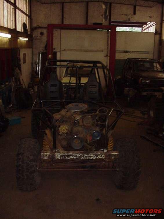 Custom buggy with exposed engine and roll cage in a workshop, another vehicle in background