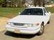 1996 Ford Crown Victoria - A white 1996 Ford Crown Victoria is parked on a paved surface with a grassy area and buildings in the background.