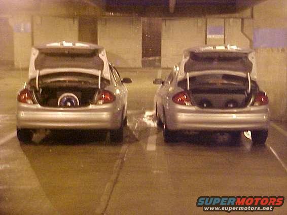 Two silver sedans parked side-by-side with open trunks, showing custom speaker setups inside