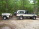 1991 Ford Bronco - A two-tone Ford Bronco tows a white camper trailer on a dirt path surrounded by trees.