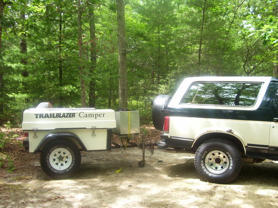 1991 Ford Bronco parked next to a Trailblazer camper trailer in a wooded area