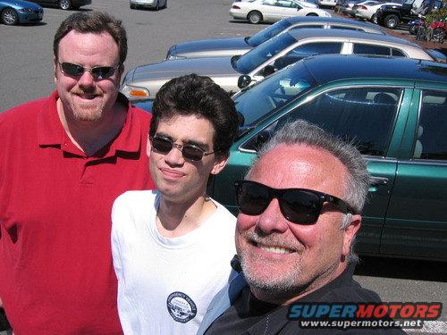 Three men pose for a selfie in front of parked cars, including a green sedan