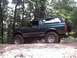 1996 Ford Bronco - A lifted, dark green Ford Bronco with off-road tires navigates a dirt path through a wooded area.