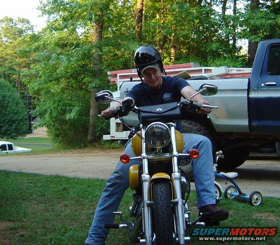 Man wearing a black helmet sits on a yellow motorcycle. A blue pickup truck is behind him