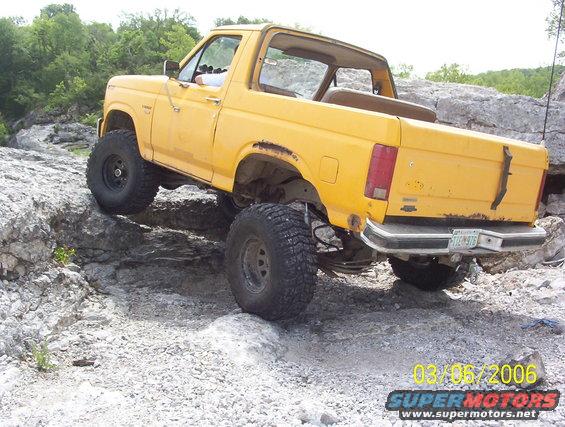 Yellow 1991 Ford Bronco climbing over rocks, rear view. License plate is visible
