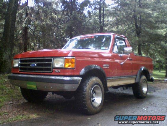 Red Ford Bronco with tan lower trim, off-road tires, parked on a dirt road in woods