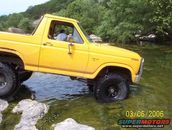 Yellow Ford Bronco driving through shallow water and over rocks. Driver visible inside