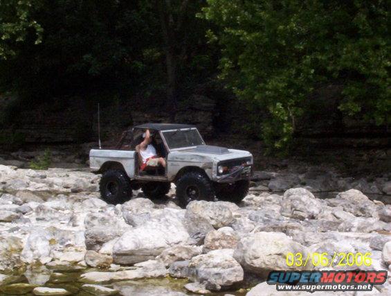 White Ford Bronco with a black top driving over rocks near trees. Driver is visible