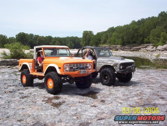Two modified Ford Broncos, one orange and one silver, parked on rocky terrain near water