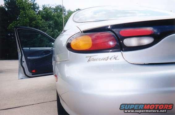 Silver sedan's rear quarter panel and open driver's door. 'Taurus GL' badge visible
