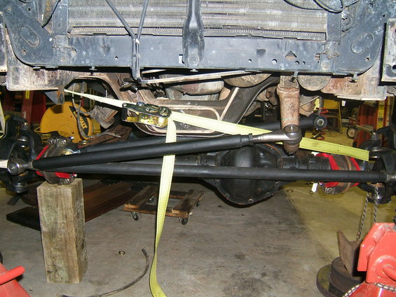 Undercarriage of a vehicle with a black steering linkage, red springs, and yellow tie-down straps. A wooden block supports the front axle