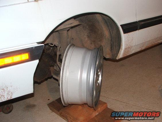 Silver aftermarket wheel resting on wooden blocks near a white car's wheel well