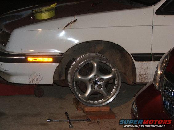 white car fender with orange turn signal, custom wheel on blocks, lug wrench on floor, red car next to it