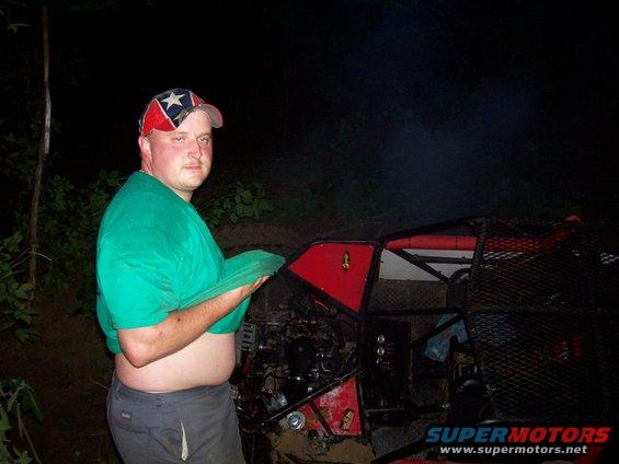 Guy in a green shirt and rebel flag hat stands next to a red off-road buggy with a roll cage. The vehicle looks modified