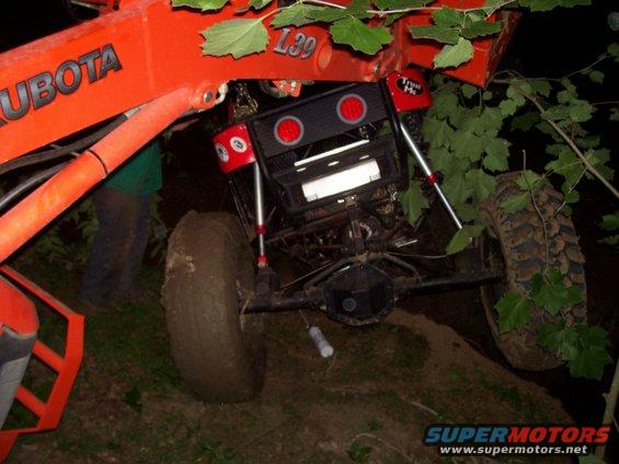 Rear view of a modified off-road vehicle with large tires, connected to a Kubota loader. A person is visible behind the loader