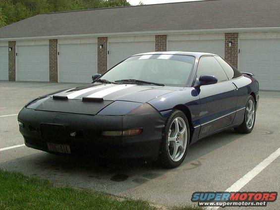Dark blue Ford Probe with white racing stripes on hood and roof, wearing a black car bra. Parked in front of a brick building with garage doors