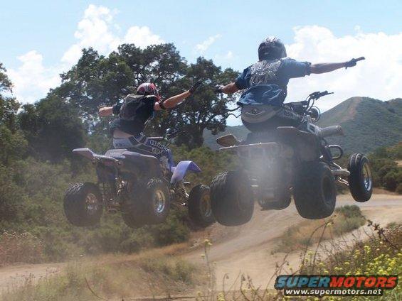 Two ATVs jump over a dirt track with riders in the air