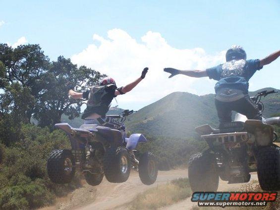 Two ATVs mid-air, riders with arms outstretched against a blue sky with clouds