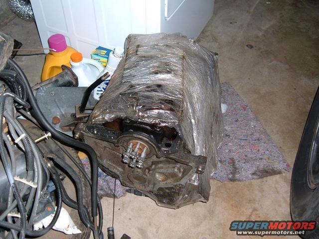 Engine block wrapped in plastic, with hoses and a tire visible