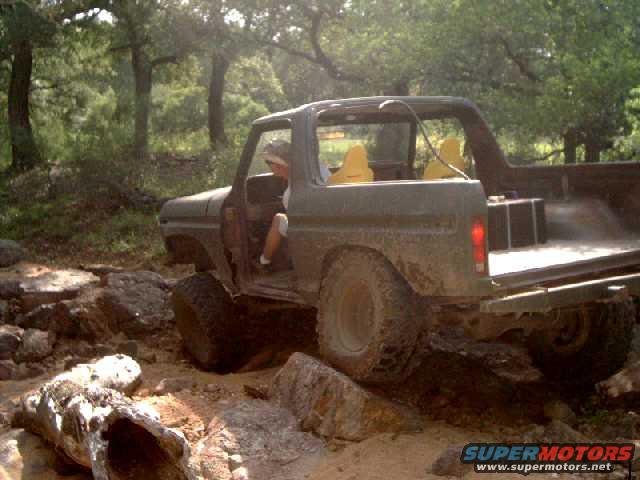 Mud-covered 1978 Ford Bronco off-roading over rocks, driver visible inside