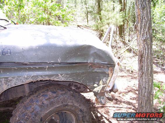 Damaged olive green vehicle fender and large mud-caked tire in wooded area. "360" is visible on the fender