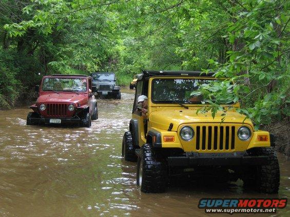 Yellow Jeep TJ front view driving in water, red Jeep TJ and gray Jeep Wrangler behind it