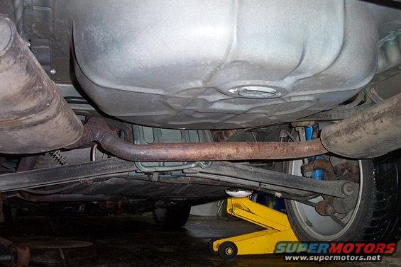 Underside view of a car's exhaust system, fuel tank, and suspension, with a yellow jack supporting the rear