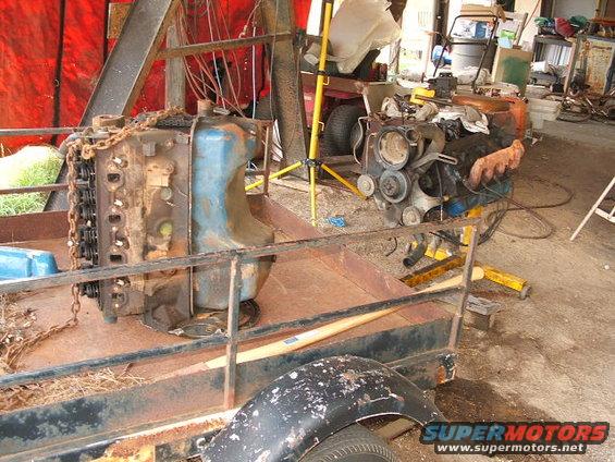Two engines on a rusty trailer in a garage. One engine is blue and black, the other is mostly black with orange exhaust manifolds