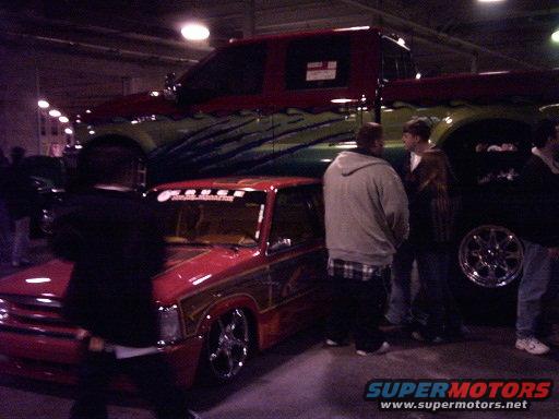 Two customized pickup trucks, one red with gold trim, the other a large red and green truck, parked indoors with people around