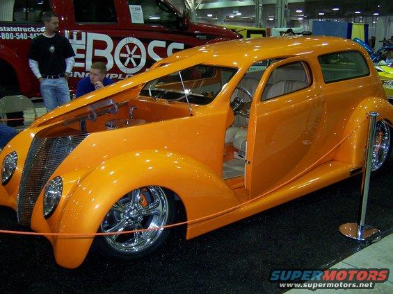 Orange custom hot rod sedan with open hood and door, chrome grille and wheels. Red truck with 'The Boss' logo behind it