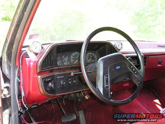 Interior shot of a vehicle's dashboard with a steering wheel, gauges, and red carpeting. Aftermarket gauges are mounted on the dash
