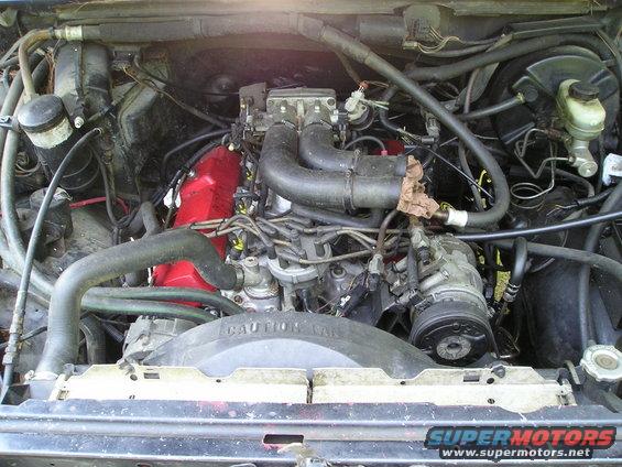 Engine bay with V8 engine, red valve covers, and fan shroud with caution text