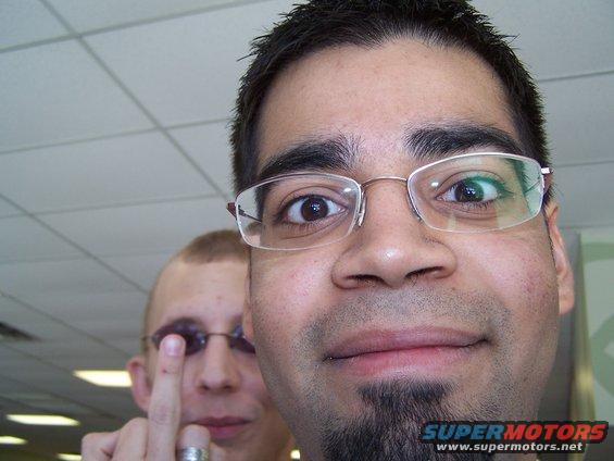 Two men posing for a selfie, one in the foreground wearing glasses and a goatee, the other in the background raising a finger