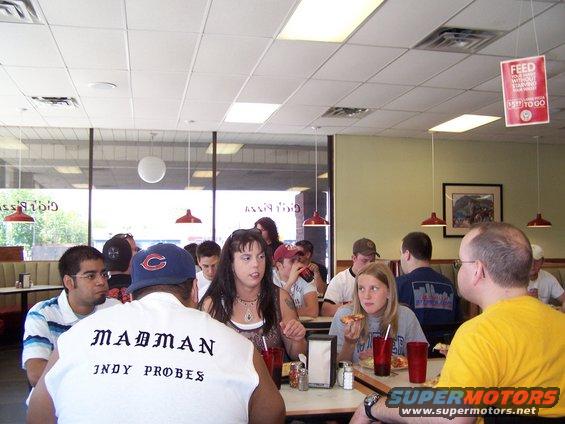 People sitting at tables in a pizza place, some eating pizza. One person wears a shirt that says "MADMAN INDY PROBES"